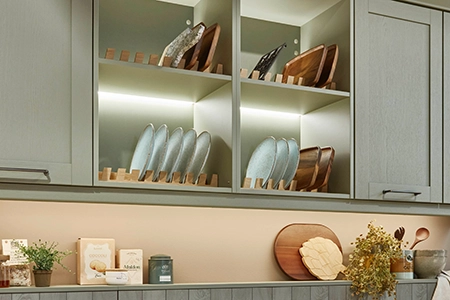 Ausschnitt einer Küche mit grünen Rahmenfronten: Wir sehen links und rechts Wandschränke, dazwischen beleuchtete Regale mit Tellereinsätzen. Ausschnitt einer Küche mit grünen Rahmenfronten: Wir sehen links und rechts Wandschränke, dazwischen beleuchtete Regale mit Tellereinsätzen.