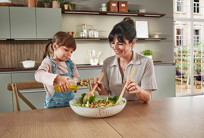 Eine Mutter sowie die etwa fünfjährige Tochter mischen Salat in einer Küche mit grünen matten Fronten. Die Mutter trägt eine beige Kurzarmbluse, die Tochter eine Latzjeans. Eine Mutter sowie die etwa fünfjährige Tochter mischen Salat in einer Küche mit grünen matten Fronten. Die Mutter trägt eine beige Kurzarmbluse, die Tochter eine Latzjeans.