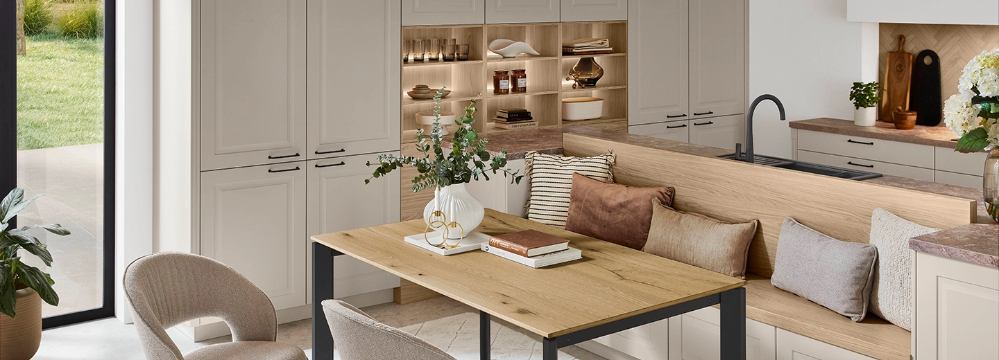 Ausschnitt einer offenen Küche im modern natural Stil. Die Rahmenfronten sind beige, im Vordergrund steht ein Esstisch mit beigen Sesseln. Ausschnitt einer offenen Küche im modern natural Stil. Die Rahmenfronten sind beige, im Vordergrund steht ein Esstisch mit beigen Sesseln.