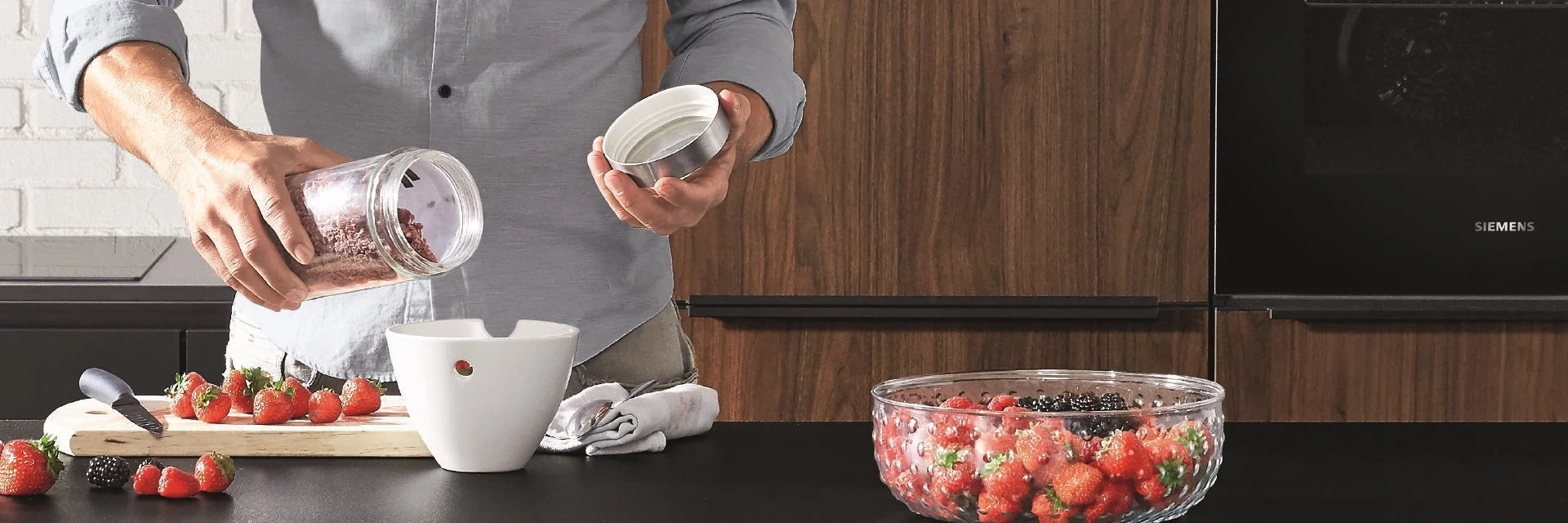 Mann in moderner Küche mit dunklen Schränken und Ziegelwand, bereitet frische Erdbeeren mit Joghurt zu Mann in moderner Küche mit dunklen Schränken und Ziegelwand, bereitet frische Erdbeeren mit Joghurt zu