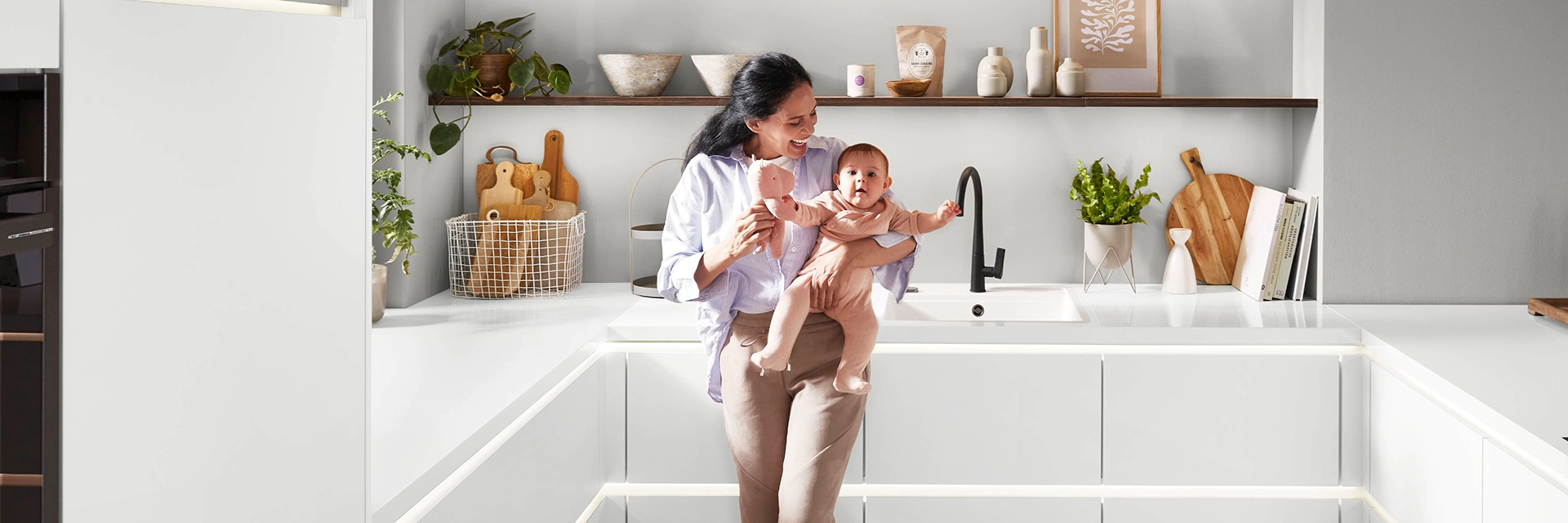 Mutter mit Baby in moderner weißer Designküche mit offenen Holzregalen und stilvoller Dekoration