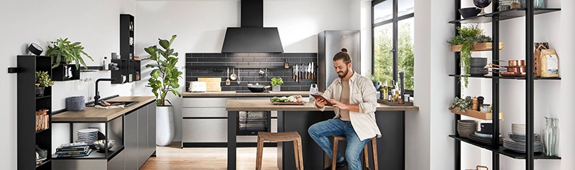Ein junger Mann sitzt in einer modernen Industrie-Stil Küche und guckt auf ein Tablet. Rechts an der Wand sind Regale mit Grünpflanzen und Dekoobjekten. Ein junger Mann sitzt in einer modernen Industrie-Stil Küche und guckt auf ein Tablet. Rechts an der Wand sind Regale mit Grünpflanzen und Dekoobjekten.