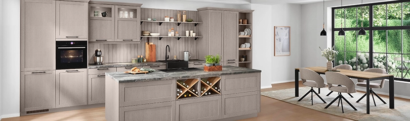 Eine moderne Küche mit einer Rahmenfront in einem hellen Kaffeeton. Sie besteht aus einer Zeile mit Hochschränken links und rechts sowie einer Kochinsel. Rechts davon ein Esstisch mit Stühlen. Eine moderne Küche mit einer Rahmenfront in einem hellen Kaffeeton. Sie besteht aus einer Zeile mit Hochschränken links und rechts sowie einer Kochinsel. Rechts davon ein Esstisch mit Stühlen.