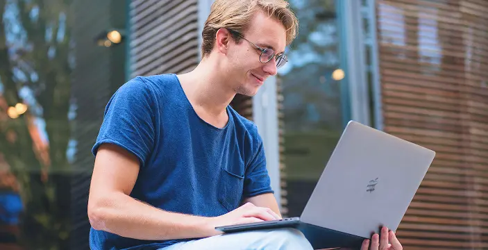 Eine Person mit blonden Haaren und Brille sitzt lächelnd im Freien und tippt auf einem silbernen Laptop. Sie trägt ein blaues Hemd und vermittelt damit eine entspannte, konzentrierte Stimmung. Eine Person mit blonden Haaren und Brille sitzt lächelnd im Freien und tippt auf einem silbernen Laptop. Sie trägt ein blaues Hemd und vermittelt damit eine entspannte, konzentrierte Stimmung.