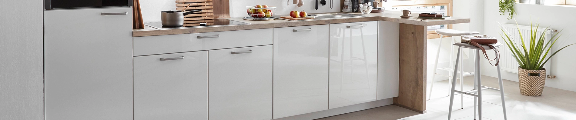 Ausschnitt einer weißen Pantryküche mit 4 Unterschränken und einem Highboard. Die Arbeitsplatte ist in einem Holzton gehalten, die Griffe sind edelstahlgrau. Ausschnitt einer weißen Pantryküche mit 4 Unterschränken und einem Highboard. Die Arbeitsplatte ist in einem Holzton gehalten, die Griffe sind edelstahlgrau.