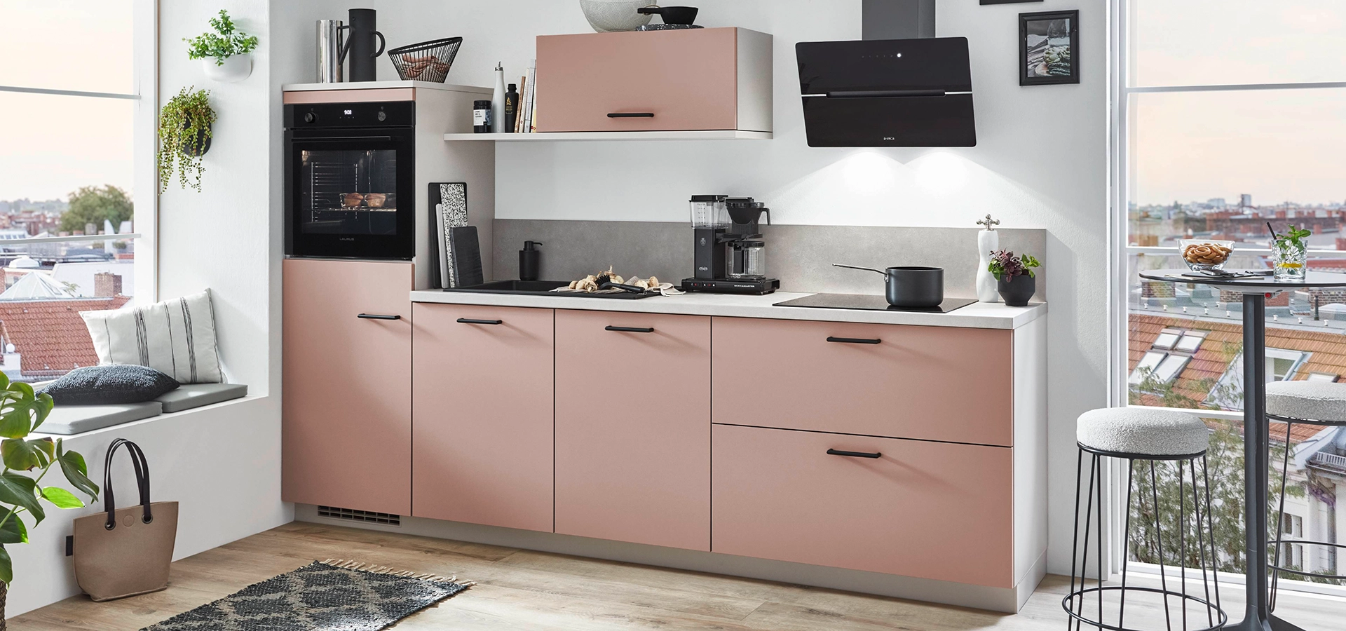 Wir sehen ein roséfarbene Pantryküche mit Highboard, schwarzer Dunsthaube und einem Wandklappenschrank. Die Griffe sind schwarz und die Arbeitsplatte weiß. Wir sehen ein roséfarbene Pantryküche mit Highboard, schwarzer Dunsthaube und einem Wandklappenschrank. Die Griffe sind schwarz und die Arbeitsplatte weiß.