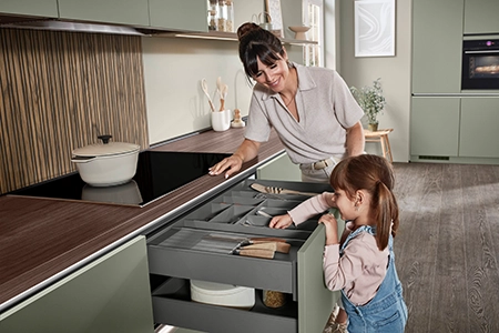 Mutter und Tochter, ca. 4 Jahre alt, stehen in einer Küche mit olivfarbenen, matten Fronten. Das Mädchen tatscht an die antifingerprintbeschichtete Front um Küchengerät aus der ausgezogenen Schublade zu holen. Mutter und Tochter, ca. 4 Jahre alt, stehen in einer Küche mit olivfarbenen, matten Fronten. Das Mädchen tatscht an die antifingerprintbeschichtete Front um Küchengerät aus der ausgezogenen Schublade zu holen.