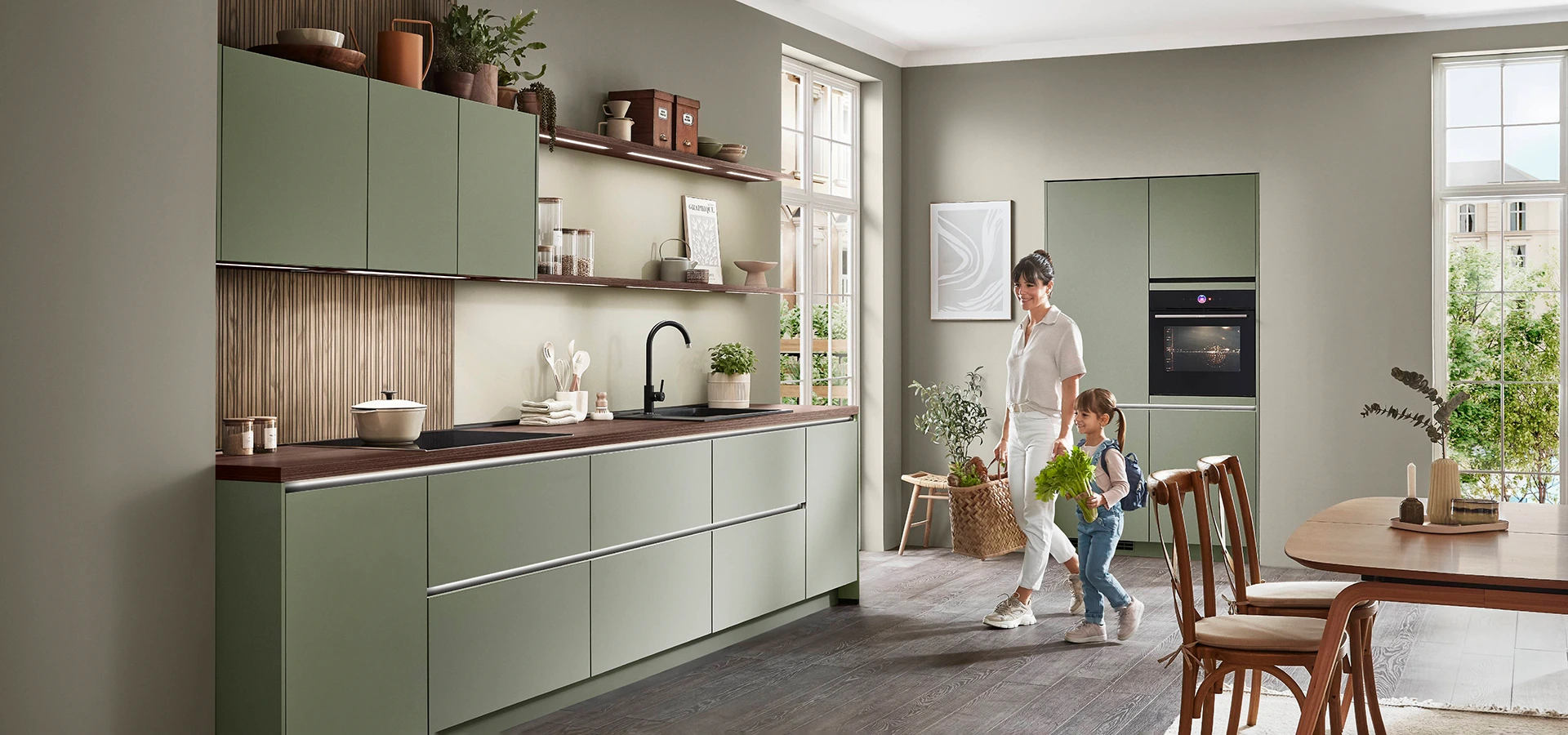 Mutter und Tochter sind in einer olivgrünen grifflosen Küche mit durchgehender Zeile sowie separat stehenden Hochschränken. Sie gehen auf die Zeile zu. Mutter und Tochter sind in einer olivgrünen grifflosen Küche mit durchgehender Zeile sowie separat stehenden Hochschränken. Sie gehen auf die Zeile zu.