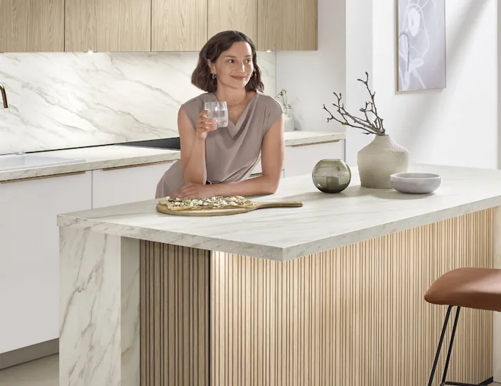 Moderne Küche mit Kochinsel in Marmoroptik und Holzlamellenfront, Frau lehnt an der Arbeitsplatte mit Getränk, helle und minimalistische Gestaltung. Moderne Küche mit Kochinsel in Marmoroptik und Holzlamellenfront, Frau lehnt an der Arbeitsplatte mit Getränk, helle und minimalistische Gestaltung.