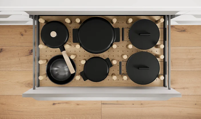 Geöffnete Küchenschublade mit organisiertem Topf- und Pfannenset in schwarzer Optik, fixiert durch variables Holz-Stecksystem auf heller Holzoberfläche. Geöffnete Küchenschublade mit organisiertem Topf- und Pfannenset in schwarzer Optik, fixiert durch variables Holz-Stecksystem auf heller Holzoberfläche.