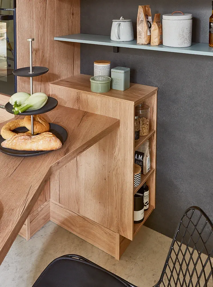 Ausschnitt einer Küche mit Holzdekor. Wir sehen den Blick auf das Ende einer Frühstückstischlösung mit anschließendem Regal. Auf dem Tisch stet eine Etagere mit Lebensmitteln, im Regal sind Gläser.