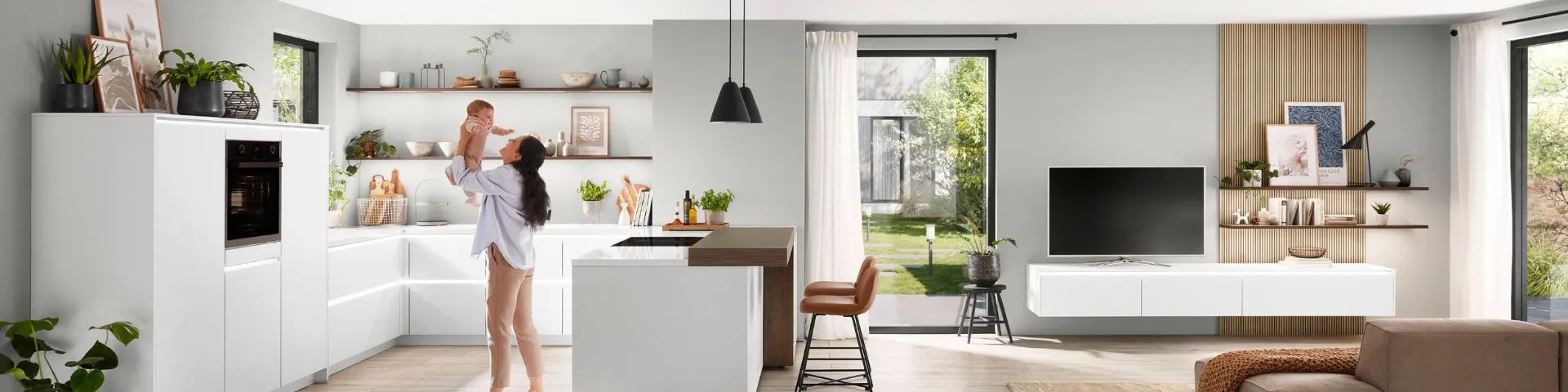 Moderne, helle Wohnküche mit weißer Einrichtung, Holzakzenten und einer Person, die ein Baby hält. Moderne, helle Wohnküche mit weißer Einrichtung, Holzakzenten und einer Person, die ein Baby hält.