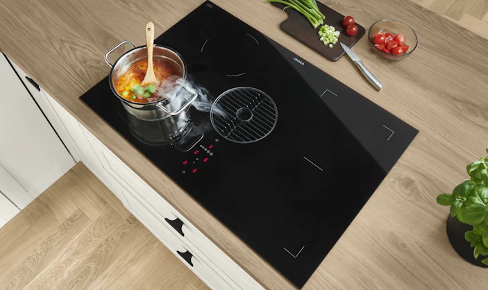 Induktionskochfeld mit integriertem Dunstabzug auf Holzarbeitsplatte, Topf mit kochender Suppe, daneben Schneidebrett mit Gemüse und Küchenutensilien. Induktionskochfeld mit integriertem Dunstabzug auf Holzarbeitsplatte, Topf mit kochender Suppe, daneben Schneidebrett mit Gemüse und Küchenutensilien.