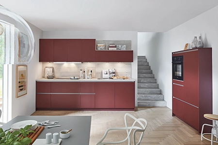 Zweizeilige Küche mit matten roten Fronten und Nischenwandbeleuchtung. Vorne links sehen wir den rechten Teil eines Esstischs sowie den oberen Teil eines Stuhls. Zweizeilige Küche mit matten roten Fronten und Nischenwandbeleuchtung. Vorne links sehen wir den rechten Teil eines Esstischs sowie den oberen Teil eines Stuhls.